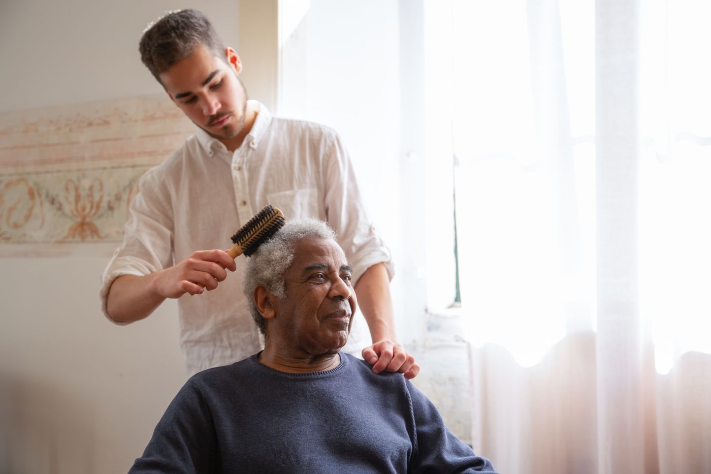 A support worker gently brushing a client's hair by a sunlit window